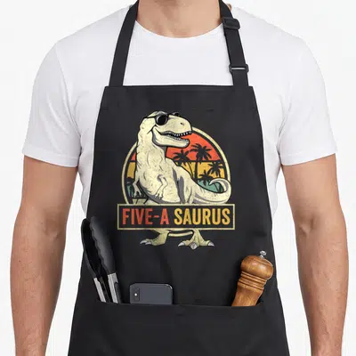 Dinosaur t-rex apron - T-Rex wearing sunglasses with FIVE-A SAURUS text over retro sunset — shown on a black cotton canvas apron against a white background
