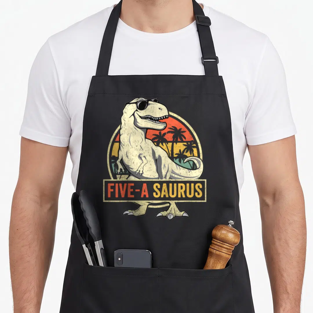 Dinosaur t-rex apron - T-Rex wearing sunglasses with FIVE-A SAURUS text over retro sunset — shown on a black cotton canvas apron against a white background