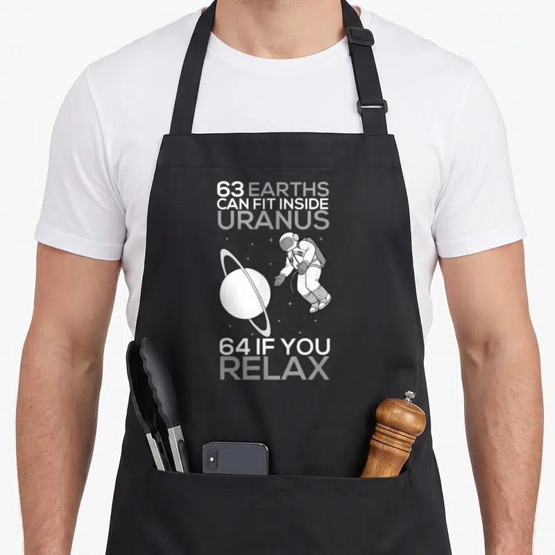 Earth quran apron - Astronaut floating near ringed planet with text 63 Earths can fit inside Uranus 64 if you relax — shown on a black cotton canvas apron against a white background
