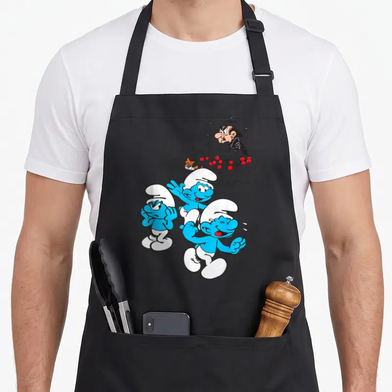 Gargamel friends cooking apron - Three blue Smurfs running with Gargamel hiding in a red berry bush background — shown on a black cotton canvas apron against a white background