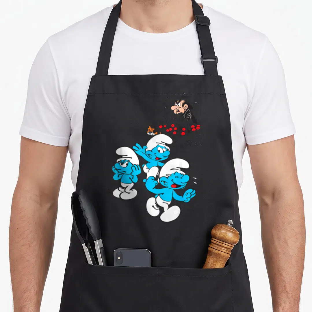 Gargamel friends cooking apron - Three blue Smurfs running with Gargamel hiding in a red berry bush background — shown on a black cotton canvas apron against a white background