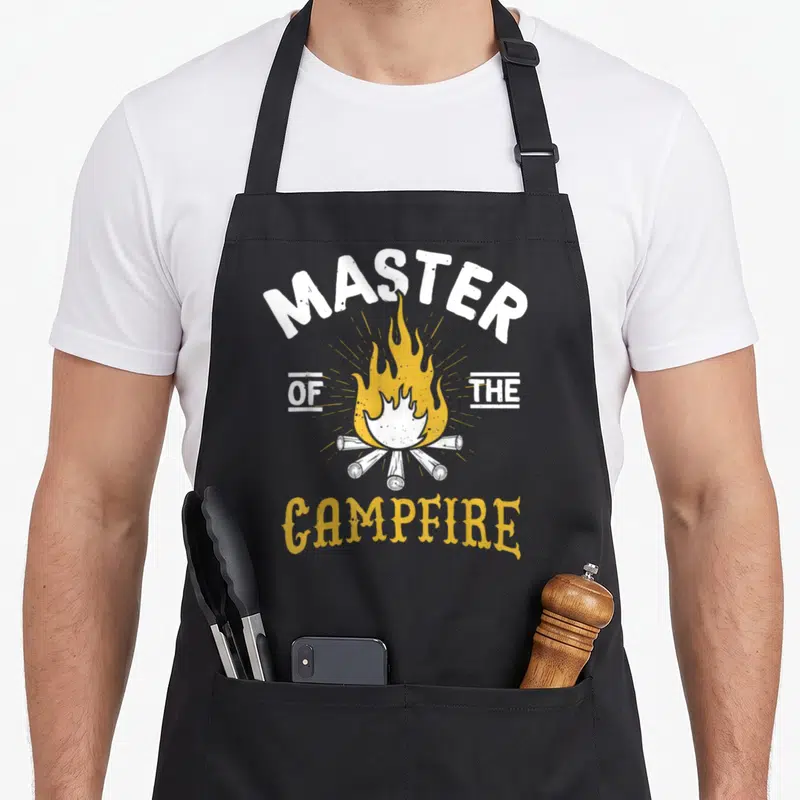 Campfire outdoor apron - White and yellow distressed campfire graphic with arched MASTER OF THE CAMPFIRE text — shown on a black cotton canvas apron against a white background