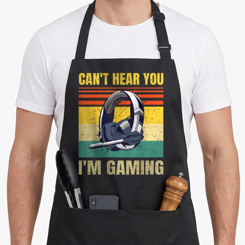 Gamer headset hear gaming apron - Retro headphone over yellow orange green blue stripes with CAN'T HEAR YOU I'M GAMING — shown on a black cotton canvas apron against a white background