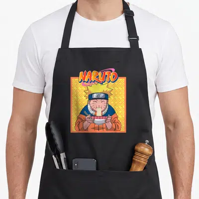 Funny eating ramen apron - Naruto cartoon character eating noodles from a bowl against yellow background with orange — shown on a black cotton canvas apron against a white background