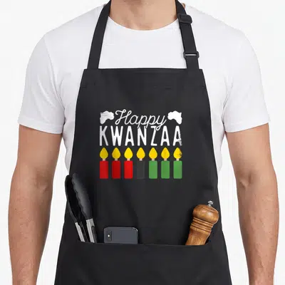Seven kwanzaa vibes apron - Happy Kwanzaa text above seven candles in red black green and yellow with Africa maps — shown on a black cotton canvas apron against a white background