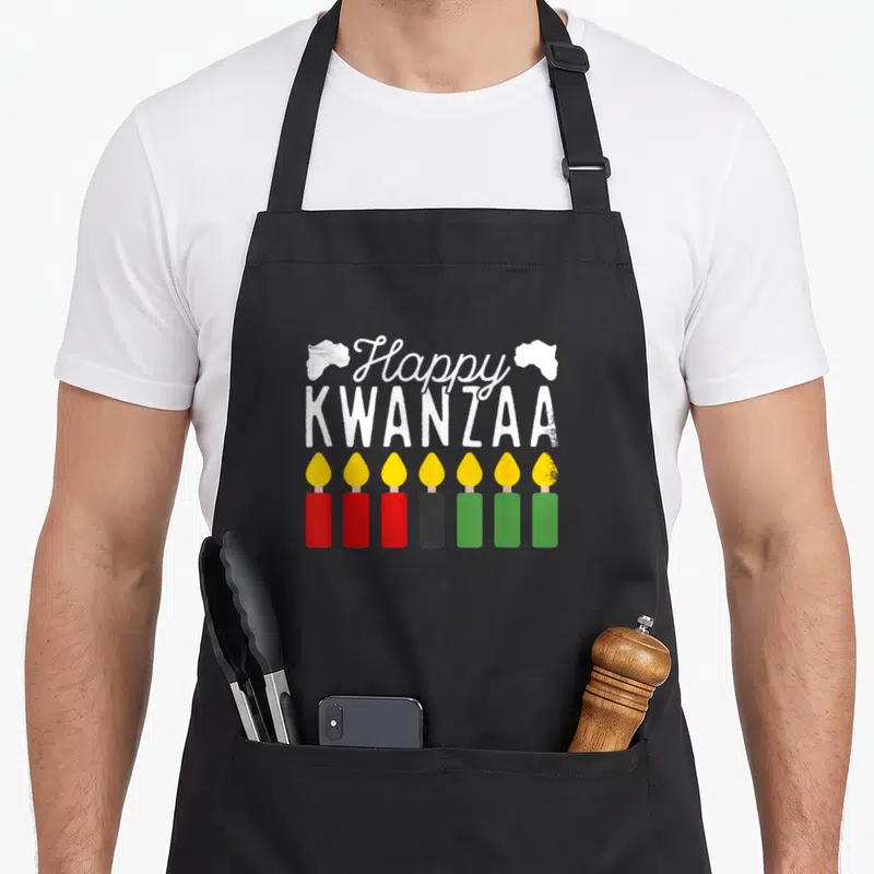 Seven kwanzaa vibes apron - Happy Kwanzaa text above seven candles in red black green and yellow with Africa maps — shown on a black cotton canvas apron against a white background