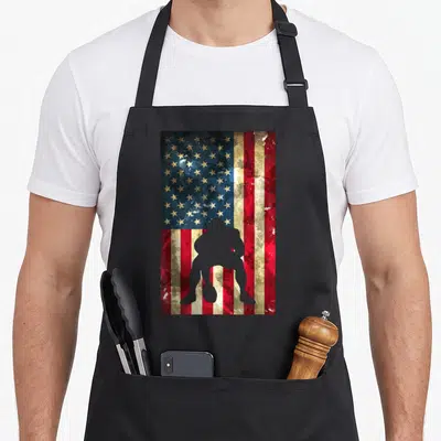 Football american flag apron - Distressed US flag with grey crouching football player silhouette, no text — shown on a black cotton canvas apron against a white background
