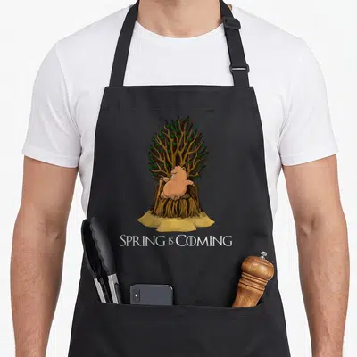 Coming spring apron - Grumpy cartoon groundhog on root throne holding cane with SPRING IS COMING text below — shown on a black cotton canvas apron against a white background