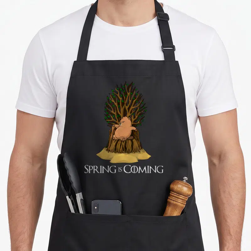 Coming spring apron - Grumpy cartoon groundhog on root throne holding cane with SPRING IS COMING text below — shown on a black cotton canvas apron against a white background