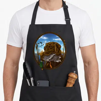 Mallard labrador apron - Brown lab holding mallard duck in marsh with blue sky and flying birds inside oval frame — shown on a black cotton canvas apron against a white background