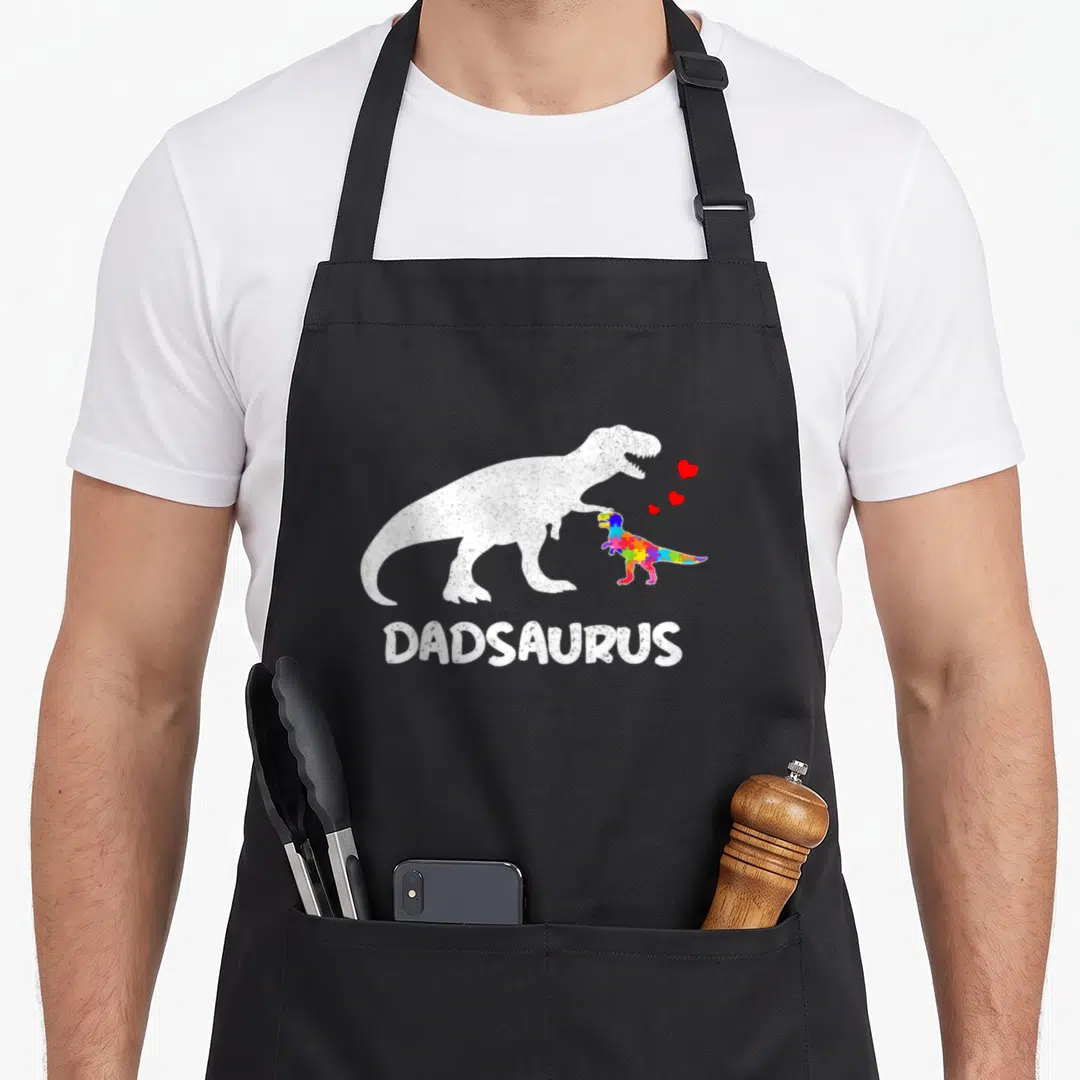 Dadsaurus dinosaur apron - White T-Rex and rainbow puzzle dinosaur with red hearts and DADSAURUS text — shown on a black cotton canvas apron against a white background