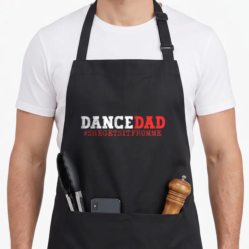 She daddy apron - DANCEDAD text in white and red with hashtag #SHEGETSITFROMME below — shown on a black cotton canvas apron against a white background