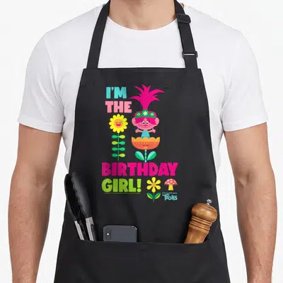 Troll birthday apron - Pink troll character with tall flower and mushroom, I'M THE BIRTHDAY GIRL! text in bright colors — shown on a black cotton canvas apron against a white background