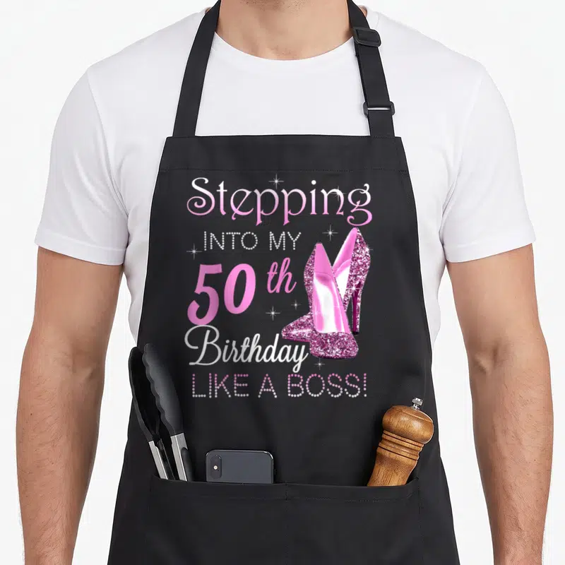 50th birthday bbq boss apron - Pink high heels and text Stepping Into My 50th Birthday Like A Boss with sparkles — shown on a black cotton canvas apron against a white background