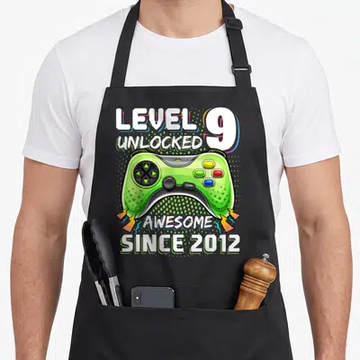 Funny level video game apron - Green game controller with LEVEL 9 UNLOCKED and AWESOME SINCE 2012 text and lightning — shown on a black cotton canvas apron against a white background