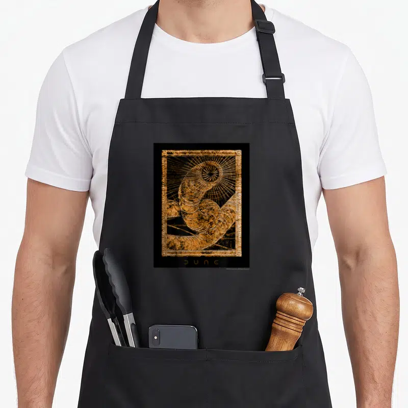 Dune golden apron - Coiled sandworm illustration in orange and black with rectangular border and centered text — shown on a black cotton canvas apron against a white background