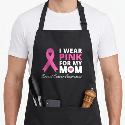 Ribbon family aprons - Pink awareness ribbon next to I WEAR PINK FOR MY MOM text with heart in M — shown on a black cotton canvas apron against a white background