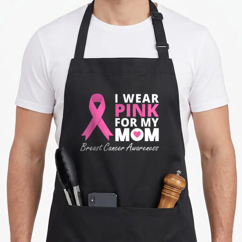 Ribbon family aprons - Pink awareness ribbon next to I WEAR PINK FOR MY MOM text with heart in M — shown on a black cotton canvas apron against a white background