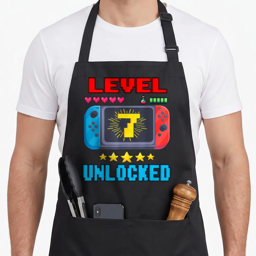 7th level birthday apron - Pixel game console with number 7 screen, LEVEL UNLOCKED text, hearts, battery, and stars — shown on a black cotton canvas apron against a white background