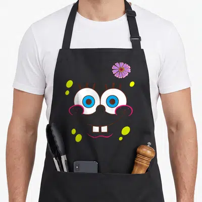 Flower face apron - Cartoon face with blue eyes, pink smile, purple flower, and yellow dots — shown on a black cotton canvas apron against a white background