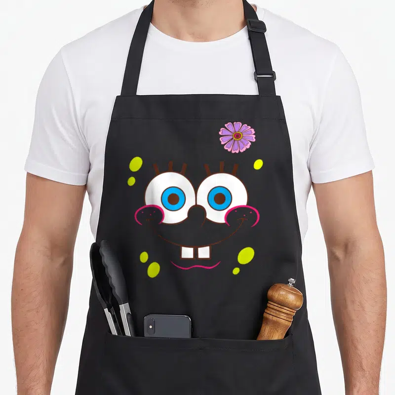 Flower face apron - Cartoon face with blue eyes, pink smile, purple flower, and yellow dots — shown on a black cotton canvas apron against a white background