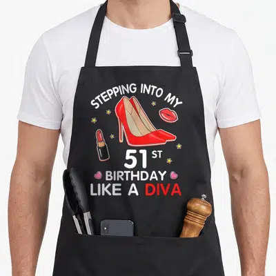 Stepping 51st birthday apron - Red high heels, lipstick, and stars surrounding 51st birthday diva text — shown on a black cotton canvas apron against a white background
