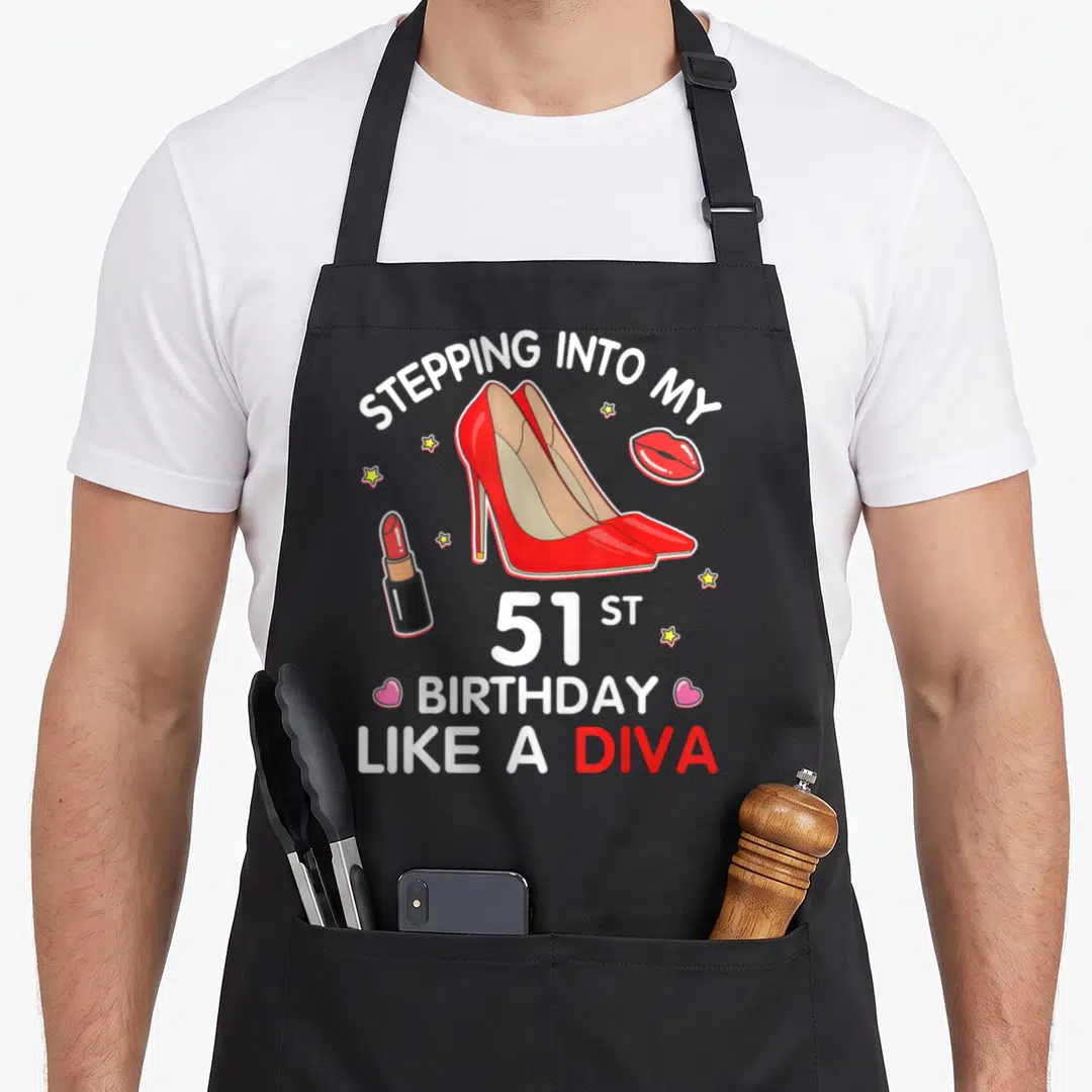 Stepping 51st birthday apron - Red high heels, lipstick, and stars surrounding 51st birthday diva text — shown on a black cotton canvas apron against a white background