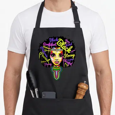 Juneteenth queen african apron - Cartoon woman face with text-filled afro in yellow purple pink green reading Strong — shown on a black cotton canvas apron against a white background