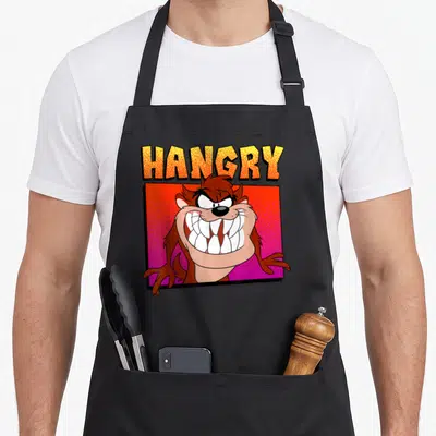 Portrait hangry apron - Taz head inside red stippled square with yellow and orange HANGRY text — shown on a black cotton canvas apron against a white background