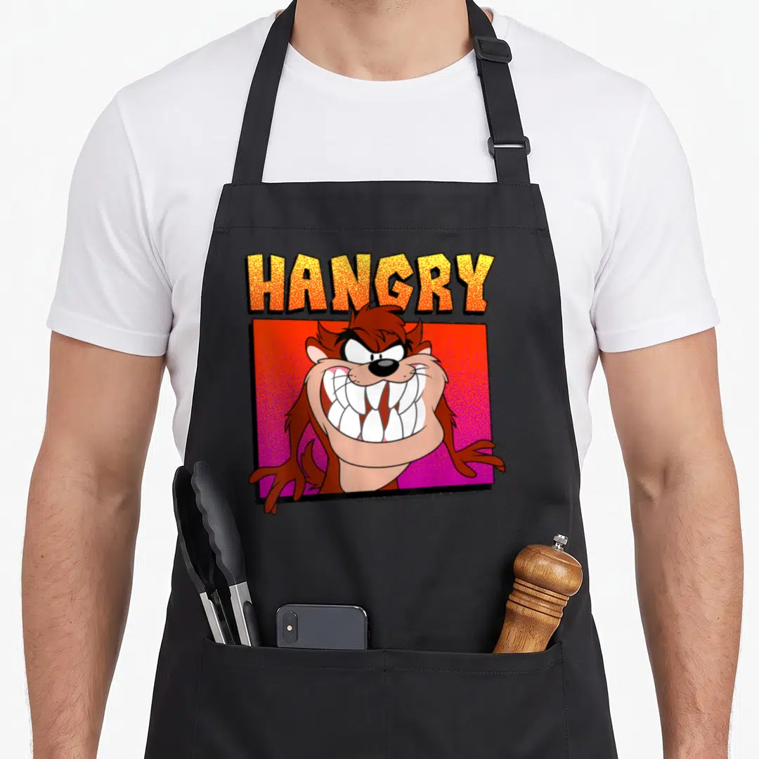 Portrait hangry apron - Taz head inside red stippled square with yellow and orange HANGRY text — shown on a black cotton canvas apron against a white background