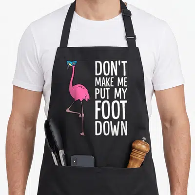 Funny awareness pink flamingo apron - Pink flamingo with blue sunglasses next to stacked white text reading DON'T MAKE — shown on a black cotton canvas apron against a white background