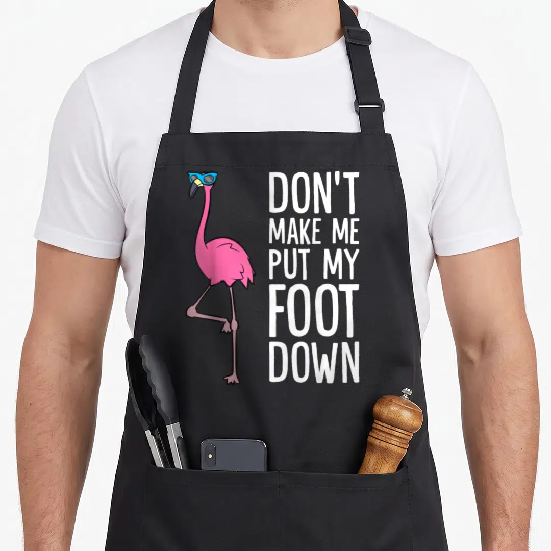 Funny awareness pink flamingo apron - Pink flamingo with blue sunglasses next to stacked white text reading DON'T MAKE — shown on a black cotton canvas apron against a white background