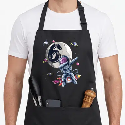 Space 6th birthday apron - Dabbing astronaut floating near number 6 and planets on grey background — shown on a black cotton canvas apron against a white background