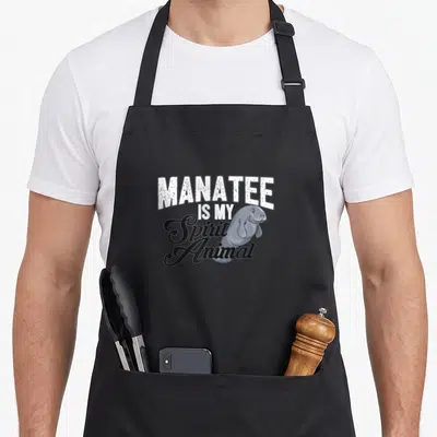 Vintage manatee apron - Cartoon manatee overlapping stacked text reading MANATEE IS MY Spirit Animal in white and black — shown on a black cotton canvas apron against a white background