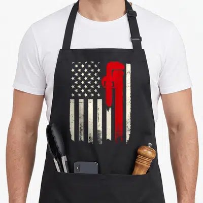 Plumbing american flag apron - Distressed US flag with large red vertical pipe wrench on right side — shown on a black cotton canvas apron against a white background