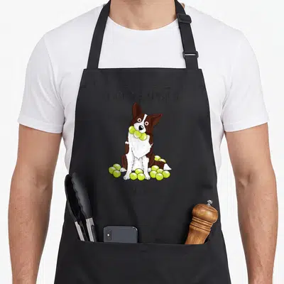 Typical border collie apron - Cartoon brown white dog holding yellow green tennis balls with obsessive pun text — shown on a black cotton canvas apron against a white background