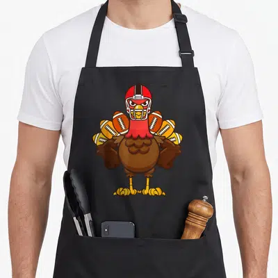 Football thanksgiving apron - Cartoon turkey in red helmet with footballs fanned as tail feathers in brown red orange — shown on a black cotton canvas apron against a white background