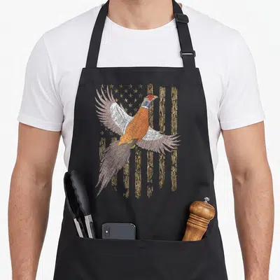 Hunting bird apron - Realistic pheasant flying over camouflage stars and stripes flag in orange brown and white — shown on a black cotton canvas apron against a white background