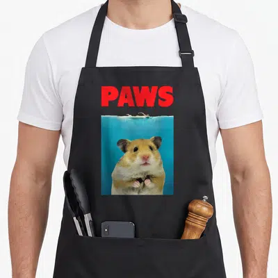 Paw hamster dad apron - Red PAWS text above a tan hamster underwater in blue water with a swimmer above — shown on a black cotton canvas apron against a white background