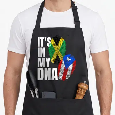 Jamaican heritage aprons - IT'S IN MY DNA text next to fingerprint flags of Jamaica and Puerto Rico — shown on a black cotton canvas apron against a white background