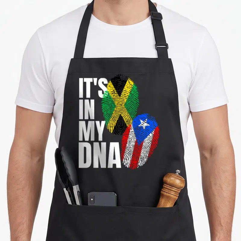 Jamaican heritage aprons - IT'S IN MY DNA text next to fingerprint flags of Jamaica and Puerto Rico — shown on a black cotton canvas apron against a white background