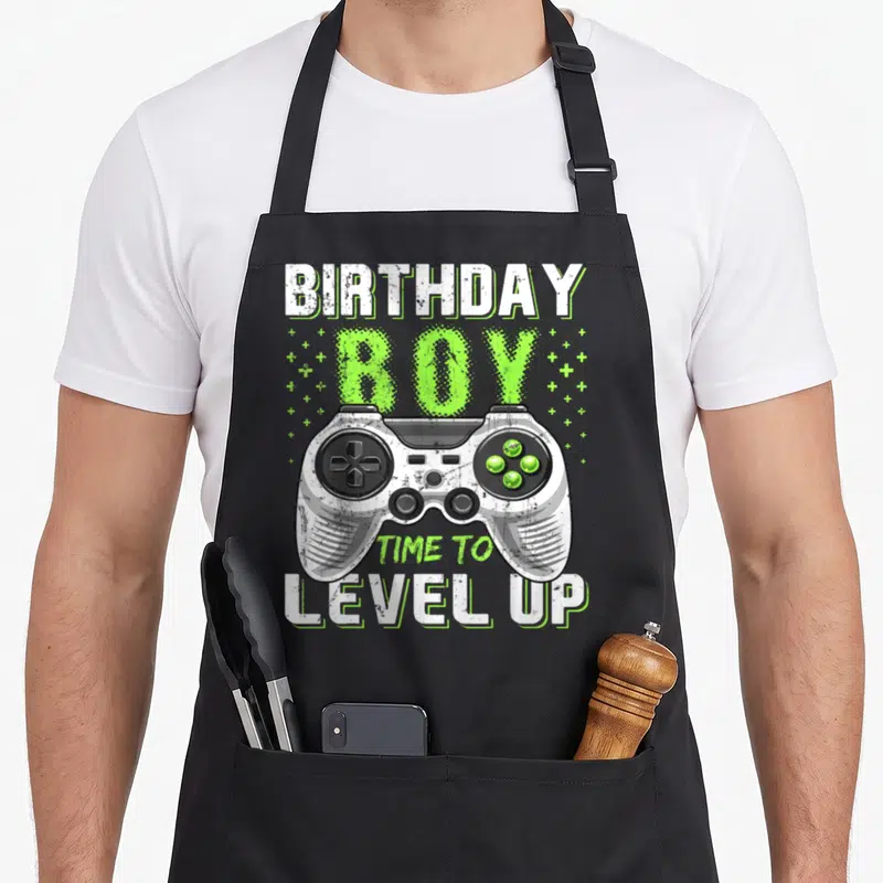 Level video game apron - Distressed controller with BIRTHDAY BOY TIME TO LEVEL UP text and plus signs — shown on a black cotton canvas apron against a white background