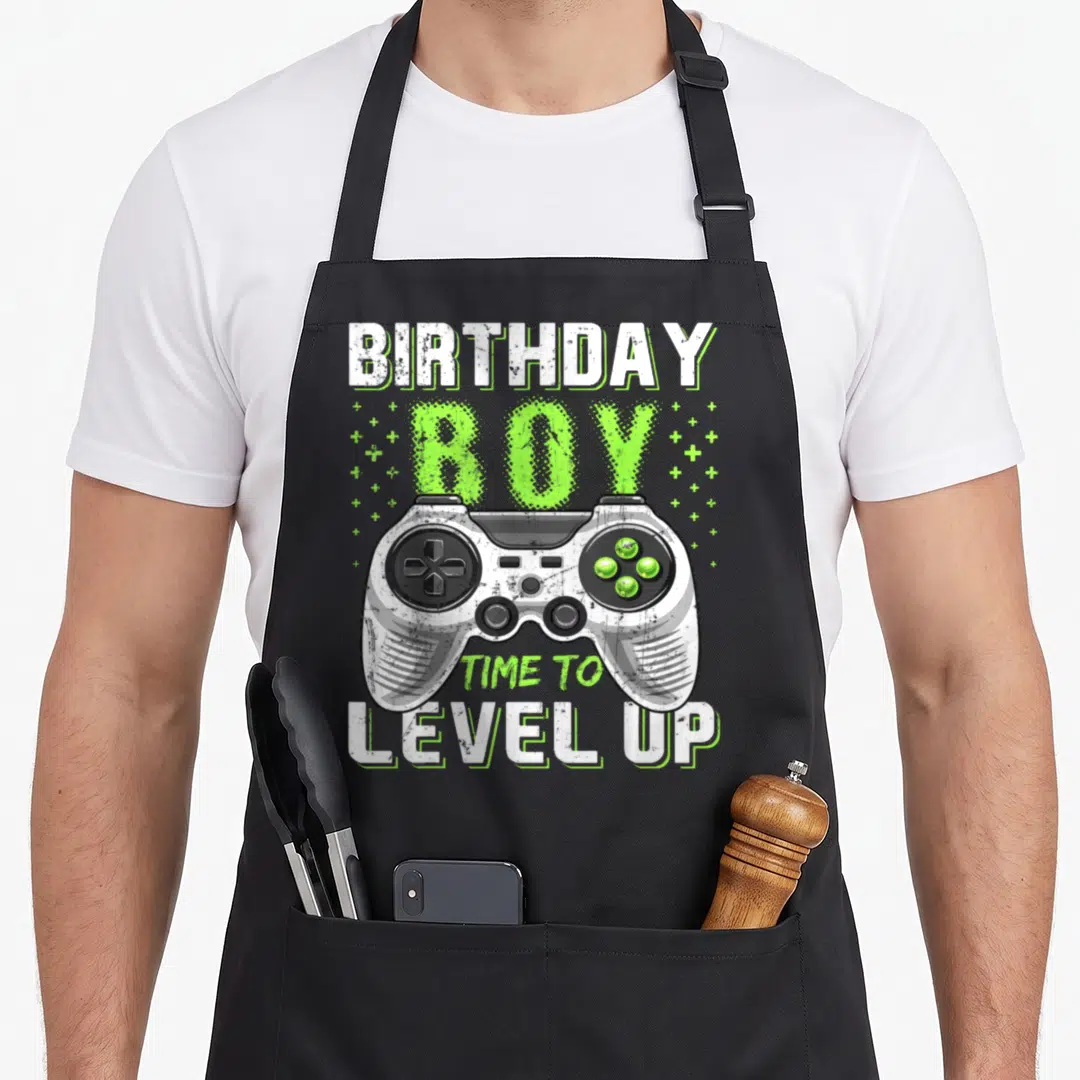 Level video game apron - Distressed controller with BIRTHDAY BOY TIME TO LEVEL UP text and plus signs — shown on a black cotton canvas apron against a white background