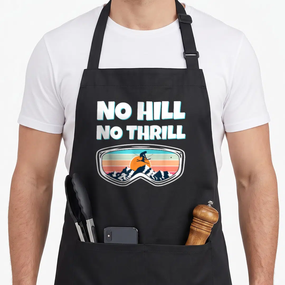 Hill goggles and apron - NO HILL NO THRILL text above ski goggles with jumping skier and sunset mountains — shown on a black cotton canvas apron against a white background