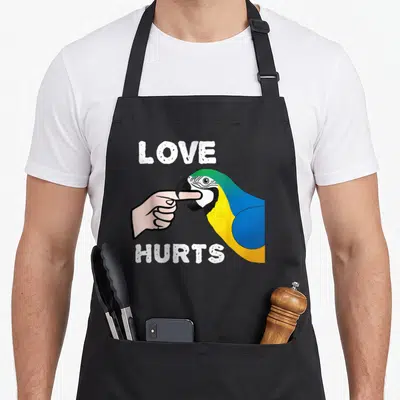 Hurt love parrot apron - Macaw biting finger between LOVE HURTS text in blue yellow green cartoon style — shown on a black cotton canvas apron against a white background