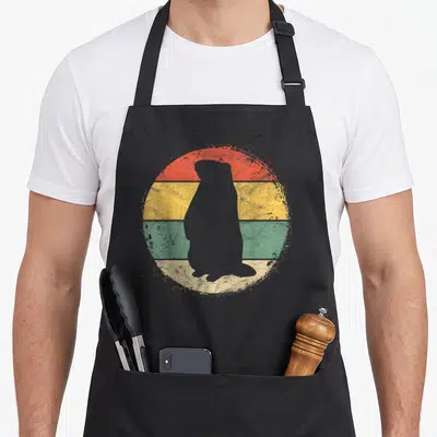 Circular groundhog apron - Grey groundhog silhouette over distressed red yellow green cream circle — shown on a black cotton canvas apron against a white background