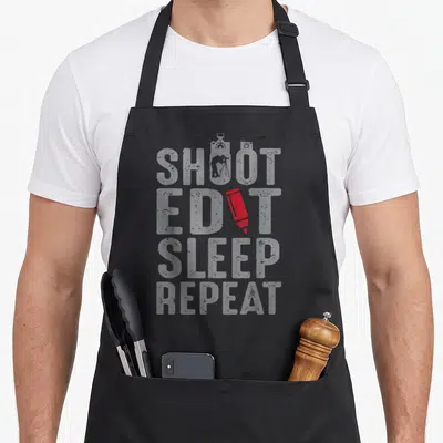 Sarcastic photographer apron - Stacked SHOOT EDIT SLEEP REPEAT text with red pencil replacing the I and camera — shown on a black cotton canvas apron against a white background