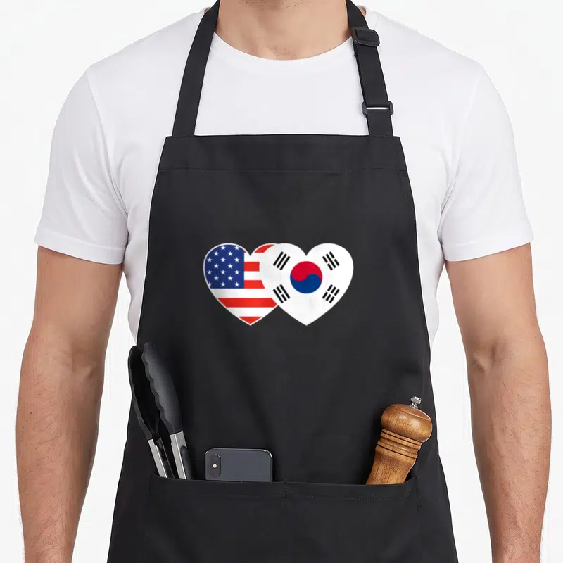 Twin american flag apron - Two overlapping heart shapes containing USA and South Korea flag patterns in red white — shown on a black cotton canvas apron against a white background