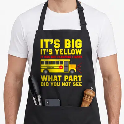 Funny school bus driver apron - Yellow school bus with red light rays and text IT'S BIG IT'S YELLOW WHAT PART DID YOU — shown on a black cotton canvas apron against a white background