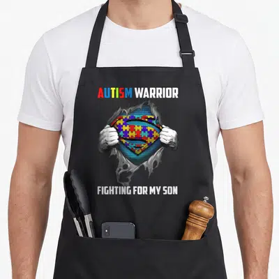 Warrior dad and son aprons - Two hands tearing grey fabric to reveal colorful puzzle piece superhero emblem with autism — shown on a black cotton canvas apron against a white background